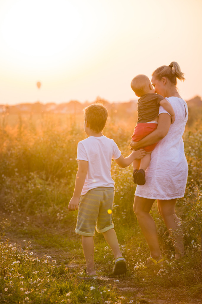 Beautiful young woman with two boys on the daisy meadow on a sunny day. Happy family on summer sunset. Kid boy giving flower to his mother. Mum with baby and kid.