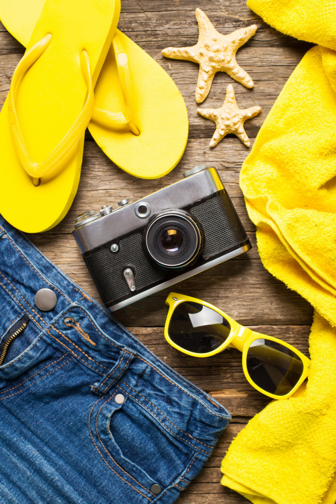 Summer women's accessories: yellow step-ins, denim shorts, camera, sunglasses on wood background.